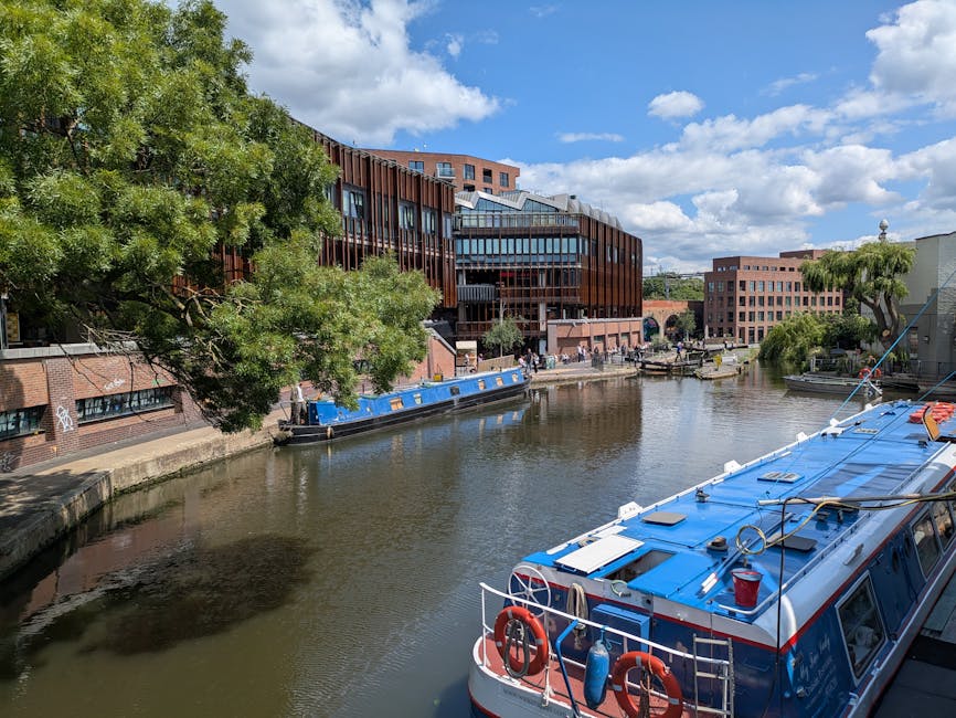 A canal scene featuring a landscaped waterside area with a mix of urban architecture and lush greenery. On the left, a large leafy tree with dense green foliage extends over the stone-paved promenade 