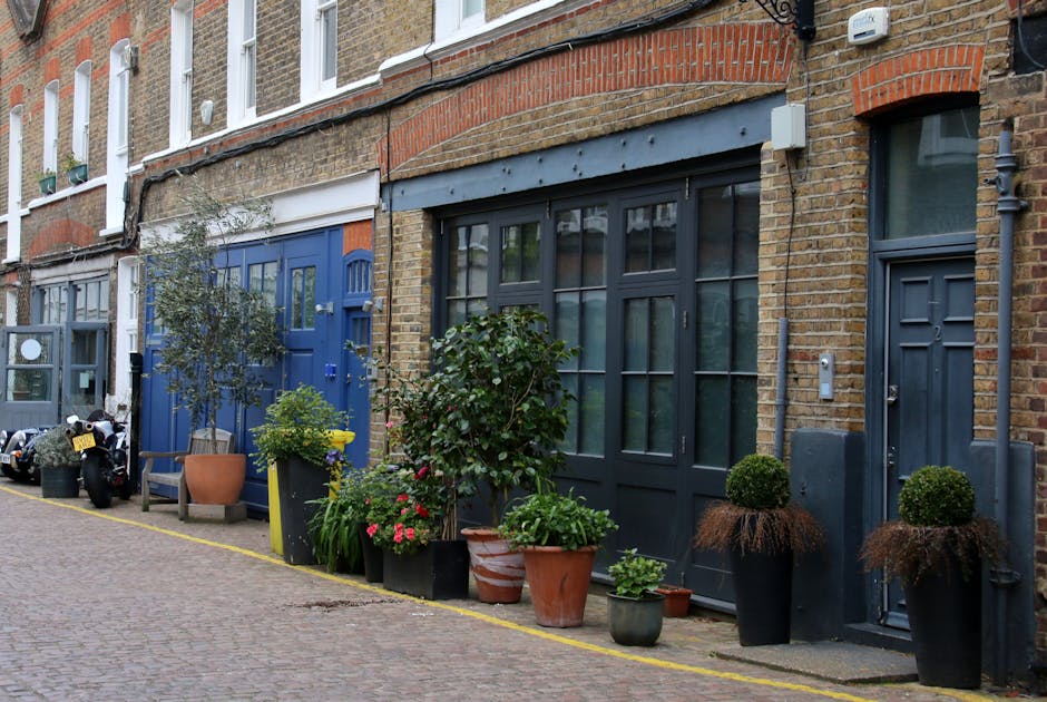 A narrow cobblestone street in a NW1 mews features a row of colorful, traditional British terraced houses with small balconies and large windows, some with black metal railings. The front gardens are